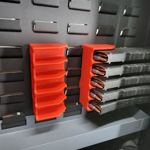 Puede incluir: Portacargadores de plástico rojo para guardar varios cargadores. Los soportes están montados en una pared de tablero perforado negro.