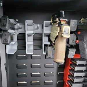 Puede incluir: Una caja fuerte para armas con una variedad de armas de fuego y cargadores almacenados en su interior. La caja fuerte tiene un interior negro y un exterior de metal gris. Las armas de fuego están dispuestas en una fila vertical, con los cargadores almacenados en una fila horizontal debajo de ellas.