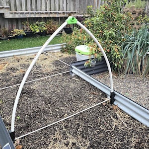 Puede incluir: Un bancal elevado con una estructura de soporte arqueada blanca. La cama está llena de tierra y paja. A la derecha hay una mesa de madera con un taladro y una valla de madera al fondo.