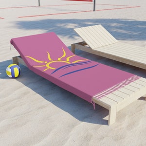 K&ouml;nnte beinhalten: Ein pinkfarbenes Strandtuch mit gelbem Sonnen- und blauem Wellenmuster, Fransen an den Kanten. Es liegt auf einer Holzliege, mit einem Volleyball im Sand. Eine weitere Liege ist im Hintergrund.