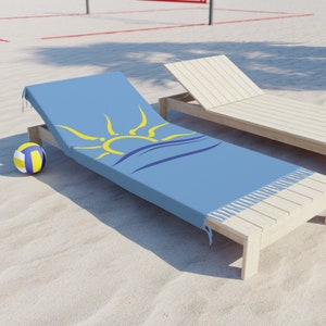 K&ouml;nnte beinhalten: Hellblaues Strandtuch mit gelbem Sonnen- und Wellenmuster, Fransen an den Kanten und einem Volleyball am Sandstrand. Im Hintergrund befindet sich ein Holz-Sonnenstuhl.