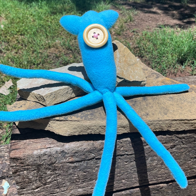 May include: A bright blue plush squid toy featuring a single large beige button sewn onto the center of its head with pink thread. The soft fabric creature has a rounded top section and several long, slender tentacles extending outward.