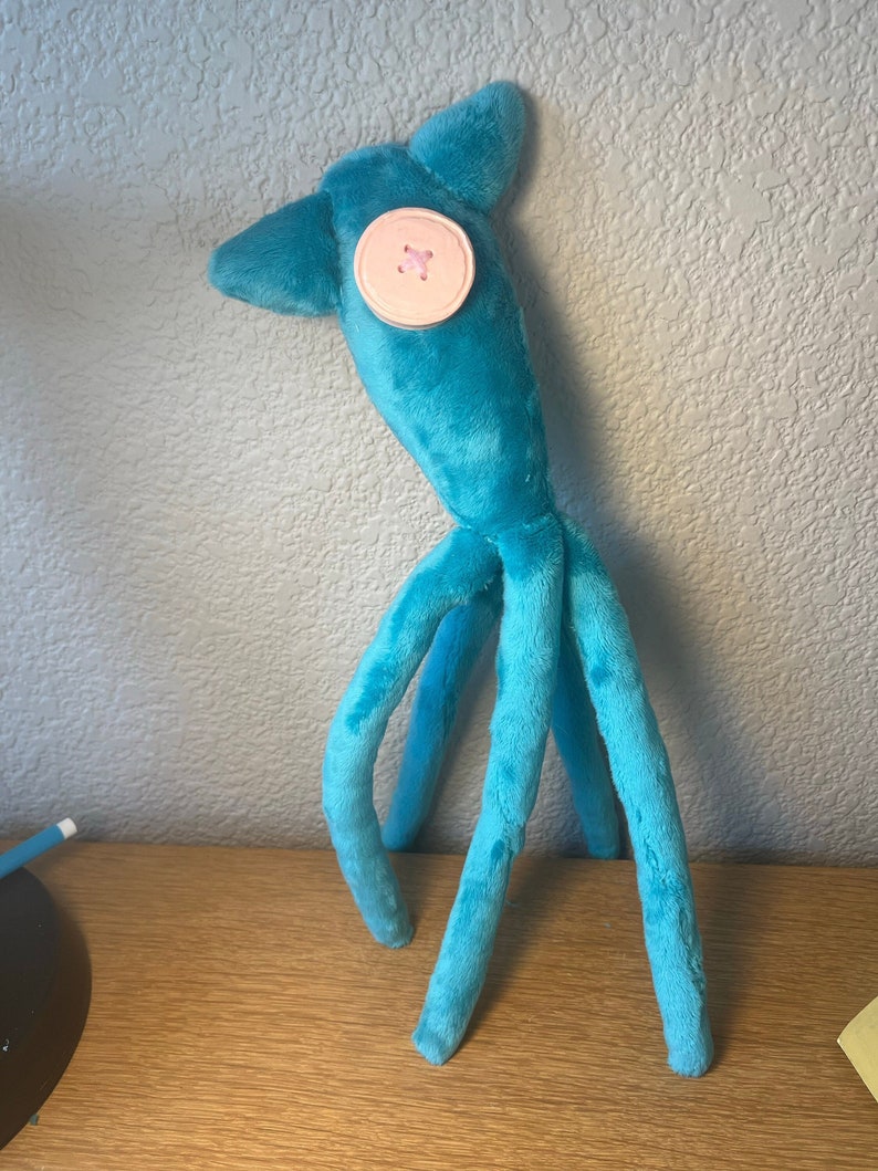 May include: A plush toy made of bright blue fabric with a soft texture. The creature has a tall, tapered head with two pointed ears and a single large, round, pale pink button sewn onto the front. It features four long, slender, dangling limbs.