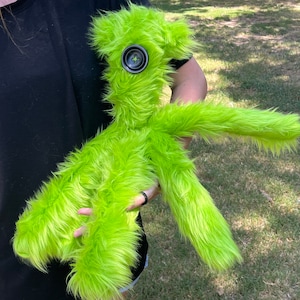 May include: A bright lime green plush toy with long, shaggy faux fur. The creature features a single large, dark gray, four-hole button centered on its face as an eye. It has a slender, elongated body with two arms and two legs, all covered in the same vibrant, textured green material.