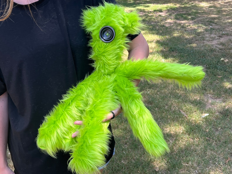 May include: A bright lime green plush toy with long, shaggy faux fur. The creature features a single large, dark gray, four-hole button centered on its face as an eye. It has a slender, elongated body with two arms and two legs, all covered in the same vibrant, textured green material.