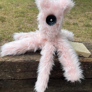 May include: A plush toy made of long, light pink faux fur with a single large black button serving as an eye. The creature features multiple long, furry limbs extending from a central body, creating a soft, textured appearance with a singular focus.