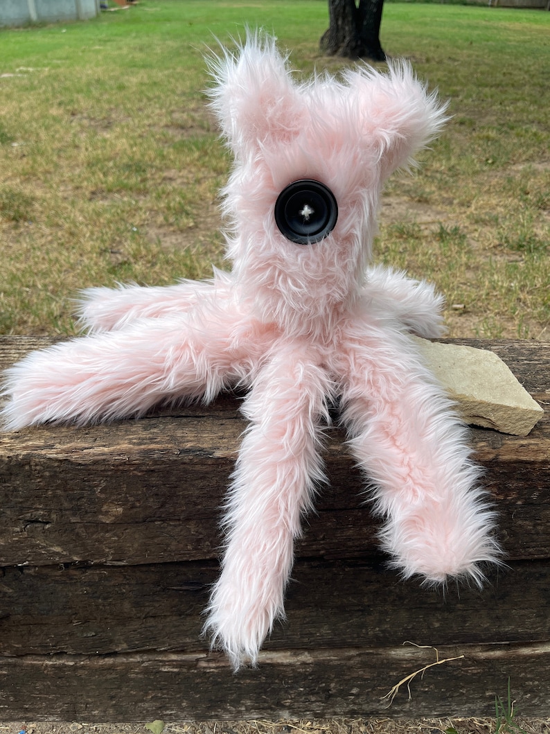 May include: A plush toy made of long, light pink faux fur with a single large black button serving as an eye. The creature features multiple long, furry limbs extending from a central body, creating a soft, textured appearance with a singular focus.