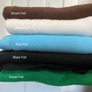 May include: A stack of five folded pieces of felt fabric in different colors. The top layer is brown, followed by white, light blue, black, and green at the bottom. Each layer is labeled with text identifying the color and material as felt.