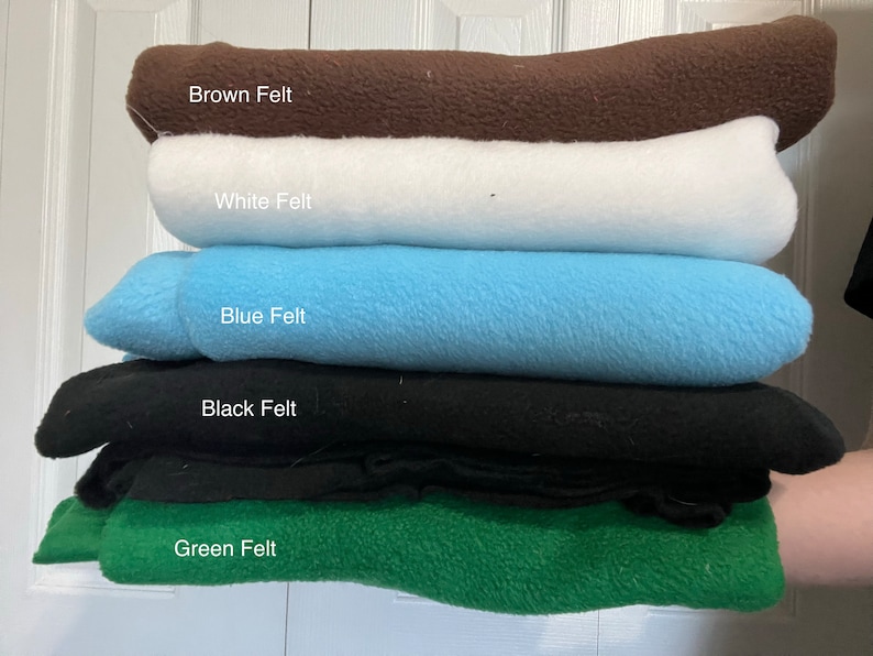 May include: A stack of five folded pieces of felt fabric in different colors. The top layer is brown, followed by white, light blue, black, and green at the bottom. Each layer is labeled with text identifying the color and material as felt.