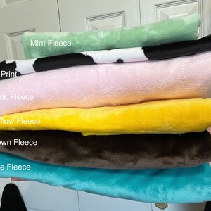 May include: A stack of six folded fleece fabric pieces in various colors and patterns. The stack includes Mint Fleece, Cow Print, Pink Fleece, Yellow Fleece, Brown Fleece, and Blue Fleece, each labeled with its respective name in white text.