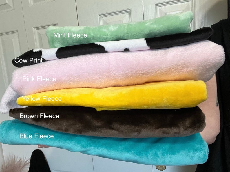 May include: A stack of six folded fleece fabric pieces in various colors and patterns. The stack includes Mint Fleece, Cow Print, Pink Fleece, Yellow Fleece, Brown Fleece, and Blue Fleece, each labeled with its respective name in white text.