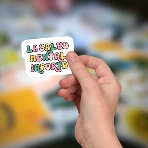 May include: A white sticker with the text "LA SALUD MENTAL IMPORTA" in colorful, retro-style lettering. The sticker is held in a hand, with a blurred background of other stickers.