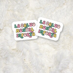 May include: Two white stickers with the text "LA SALUD MENTAL IMPORTA" in a retro font, featuring pink, yellow, and green colors. The stickers are on a fluffy white background.