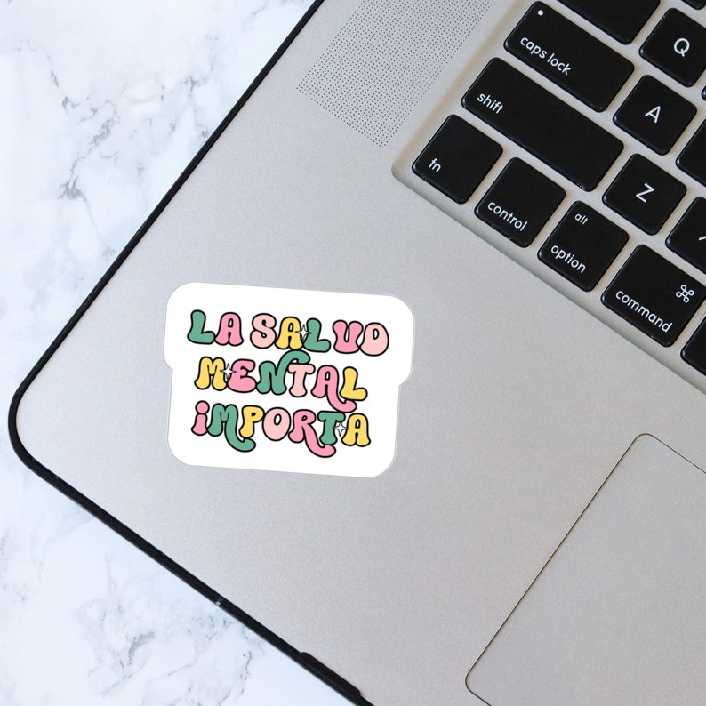 May include: A white sticker with the text "LA SALUD MENTAL IMPORTA" in a retro font. The text is in green, yellow, and pink. The sticker is on a silver laptop.
