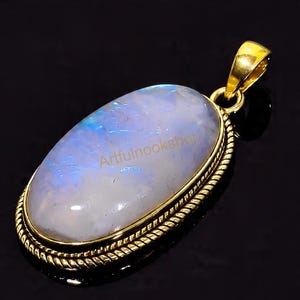 May include: Oval moonstone pendant with a gold-colored setting. The moonstone has a pearly white and blue iridescence. The pendant has a decorative rope-like border and a gold bail. The text "Artfulnookshop" is visible on the moonstone.