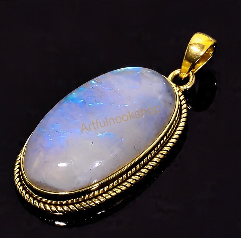 May include: Oval moonstone pendant with a gold-colored setting. The moonstone has a pearly white and blue iridescence. The pendant has a decorative rope-like border and a gold bail. The text "Artfulnookshop" is visible on the moonstone.