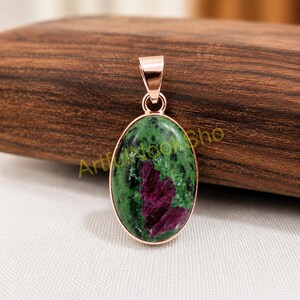 May include: An oval pendant featuring a polished zoisite stone with deep green and black inclusions and a prominent magenta-coloured ruby centre. The stone is set in a smooth rose gold-toned metal frame with a matching bail at the top.