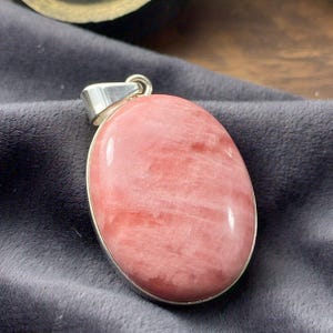 Peut inclure: Un pendentif en argent avec une grande pierre précieuse rose de forme ovale, à la surface lisse et polie. La pierre précieuse présente un motif strié subtil.