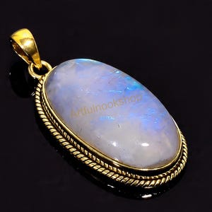 May include: A gold-colored pendant with an oval, iridescent moonstone. The moonstone has a pearly white and blue hue. The pendant is framed with a gold-colored, rope-like border. The text "Artfulnookshop" is visible on the moonstone.