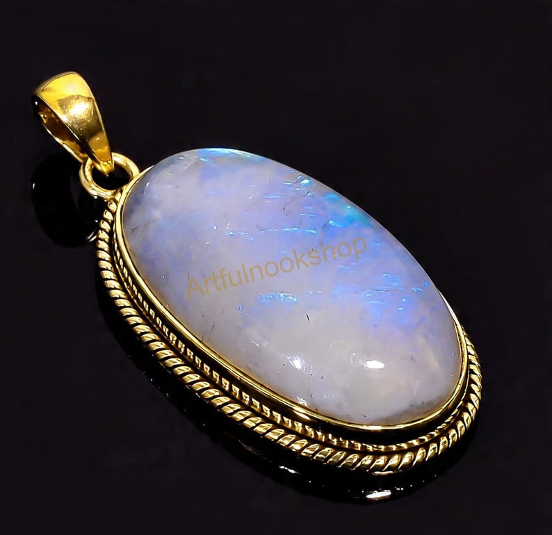 May include: A gold-colored pendant with an oval, iridescent moonstone. The moonstone has a pearly white and blue hue. The pendant is framed with a gold-colored, rope-like border. The text "Artfulnookshop" is visible on the moonstone.
