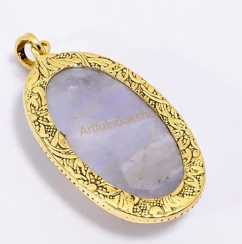 May include: Oval-shaped pendant with a gold-colored frame and a light blue stone center. The frame features an ornate, floral design. The text "Artfulnooksho" is visible on the stone. The pendant has a loop for a chain.