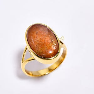 Puede incluir: Un anillo dorado con una piedra preciosa ovalada de color marrón anaranjado. El anillo tiene un diseño simple y elegante con una banda lisa y pulida y un engaste de bisel. La piedra preciosa tiene un aspecto moteado.