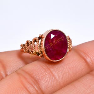 Op de afbeelding: Een roségouden ring met een grote, ovaal geslepen robijn. De ring heeft een touwachtig ontwerp op de band en de tekst "ookshop" is zichtbaar op de edelsteen. De robijn heeft een dieprode kleur.