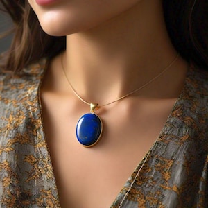 Puede incluir: Un collar dorado con un colgante grande, ovalado y azul oscuro. El colgante está engastado en un marco dorado y cuelga de una delicada cadena dorada. El collar lo lleva una persona que lleva una blusa estampada.