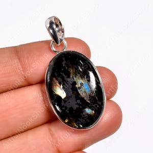 Pode incluir: Pingente oval com uma pedra preta, com flashes iridescentes azuis e amarelos. A pedra é colocada numa moldura de metal prateado com uma argola para uma corrente. Uma joia única.
