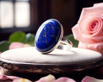 May include: A silver ring with a large oval lapis lazuli gemstone. The ring is set on a white display stand with pink rose petals scattered around it.