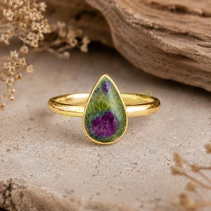 Puede incluir: Un anillo de oro con una piedra preciosa de rubí zoisita en forma de lágrima. La piedra muestra una matriz moteada verde y negra con prominentes inclusiones de color magenta intenso. El aro de metal es liso, pulido y circular, con la piedra en engaste.