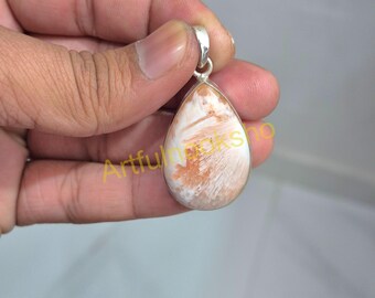 Op de afbeelding: Een druppelvormige hanger met een zilveren lus. De hanger heeft een crèmekleurige steen met bruine en witte adering. De steen is gezet in een zilveren bezel. De hanger wordt in een hand gehouden.