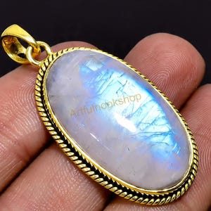 May include: Oval-shaped pendant featuring a large, iridescent moonstone with blue and white hues. The stone is set in a gold-colored metal frame with a rope-like border. The text "Artfulnookshop" is visible on the stone.