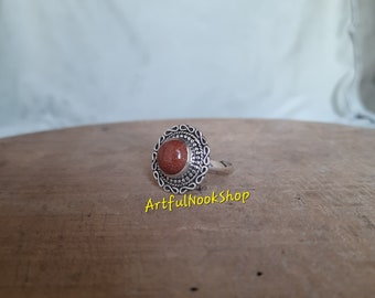 May include: A silver ring with a brown stone set in a decorative filigree design. The ring is on a wooden surface.
