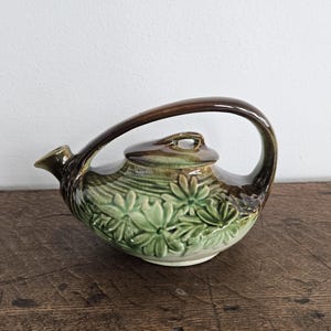 May include: A green and brown ceramic teapot with a floral design. The teapot has a curved handle and a lid with a knob.
