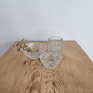 May include: Three clear glass bowls with a diamond pattern on a wooden surface. The bowls are different sizes and are arranged in a row. A small bouquet of pink flowers is in the largest bowl.