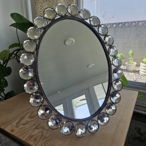 May include: Oval mirror with a dark metal frame and a border of large, clear, faceted glass stones. The mirror is set on a light-colored wooden surface, with greenery visible in the background. The mirror reflects the interior of a room.