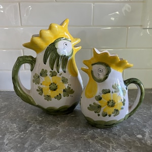 May include: Two ceramic rooster-shaped pitchers. The pitchers are white with yellow crests, green handles, and floral designs. The larger pitcher is approximately 10 inches tall, and the smaller one is about 6 inches tall. The pitchers are on a gray surface.