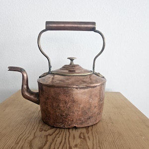 May include: A vintage copper kettle with a tarnished finish and a wooden handle. The kettle has a rounded body and a spout on the side.