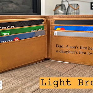 May include: Open light brown leather wallet with multiple card slots, filled with various credit and identification cards. The wallet has the text "Dad: A son's first hero, a daughter's first love." The wallet is open, displaying its contents.
