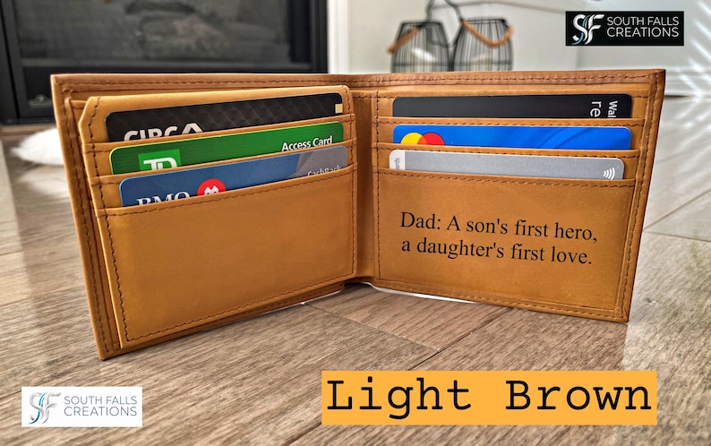 May include: Open light brown leather wallet with multiple card slots, filled with various credit and identification cards. The wallet has the text "Dad: A son's first hero, a daughter's first love." The wallet is open, displaying its contents.