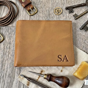 May include: A tan leather wallet with the initials "SA" embossed on the front. The wallet is surrounded by leather belts, antique keys, and tools, suggesting a handcrafted theme. The wallet is approximately 4.5 inches wide and 3.5 inches tall.