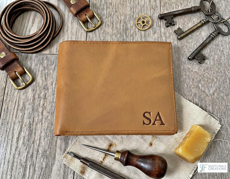 May include: A tan leather wallet with the initials "SA" embossed on the front. The wallet is surrounded by leather belts, antique keys, and tools, suggesting a handcrafted theme. The wallet is approximately 4.5 inches wide and 3.5 inches tall.