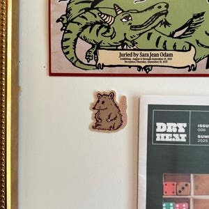 Może przedstawiać: Brązowa naklejka z kreskówkowym szczurem z różowym ogonem na białej ścianie. Nad naklejką plakat z zieloną ilustracją smoka i napisem "full on fantasy". Poniżej naklejki magazyn z napisem "DRY HEAT".