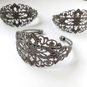 May include: Three dark gray filigree metal cuff bracelets. The bracelets feature an openwork floral design with intricate details. The bracelets are displayed on a white surface, showcasing their ornate patterns and vintage style.