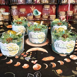 Peut inclure: Plusieurs bougies en verre avec de la cire turquoise et des garnitures décoratives sont présentées sur une surface noire avec un motif céleste. Les bougies ont des étiquettes avec du texte français. Des étagères de petits bocaux en verre remplis d'articles sont en arrière-plan.