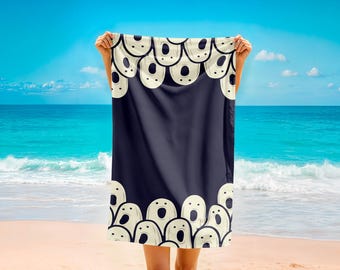 Può includere: Un telo mare blu navy con un motivo ripetuto di facce bianco sporco a bocca aperta lungo i bordi. L'asciugamano è tenuto contro uno sfondo di spiaggia sabbiosa, oceano e cielo blu.