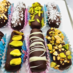 May include: Close-up of various chocolate-covered sweets in patterned paper cases. The dark chocolate treats are decorated with nuts, coconut flakes, pistachios, and white chocolate. The paper cases feature floral and geometric designs.