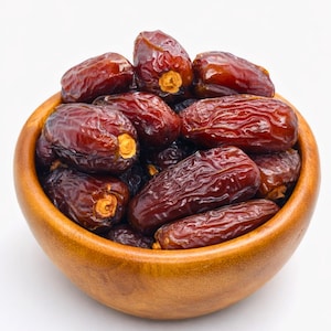 May include: A wooden bowl filled with plump, dark brown dates. The dates have a wrinkled texture and a glossy sheen, with a few showing their light-colored stems. The bowl is made of light-colored wood.