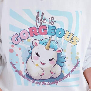 Puede incluir: Sudadera blanca con un gráfico colorido de un unicornio de dibujos animados y las palabras "Life is Gorgeous". El unicornio tiene una melena y un cuerno azules. El fondo tiene rayas azules y blancas y las palabras "when you choose to see the beauty in every moment."
